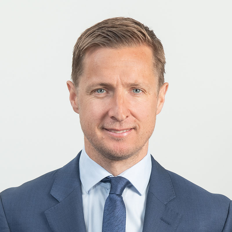 Headshot of a business executive photographed looking at the camera against a white wall. The portrait has a feel of a studio shoot through the use of professional lighting.